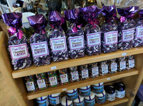 Bags of purple-wrapped chocolate almonds displayed on a wooden shelf, alongside various colorful candies and jars.