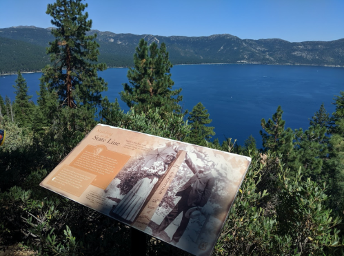 A scenic view of a lake with mountains in the background, featuring a historical sign and surrounding trees.