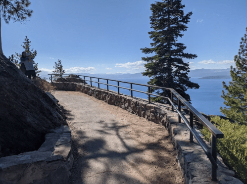 A scenic pathway with a railing, surrounded by trees, overlooking a lake and distant mountains under a clear blue sky.
