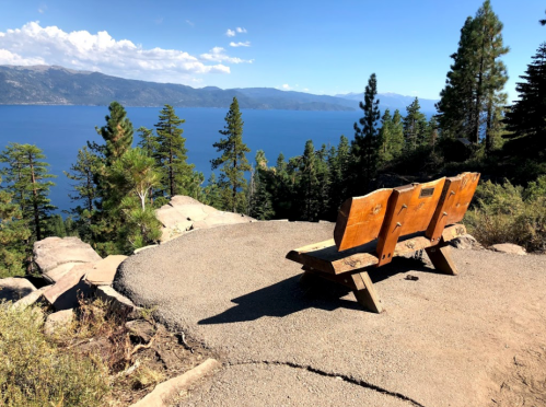 A wooden bench overlooks a serene lake surrounded by trees and mountains under a clear blue sky.