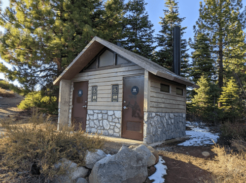 A rustic restroom facility with two doors, surrounded by trees and snow, set in a natural outdoor environment.