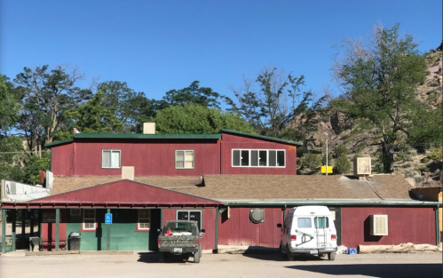 A red two-story building with a green porch, surrounded by trees and a parked RV in front. Clear blue sky above.