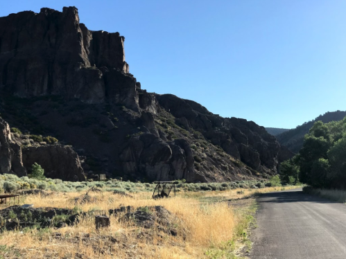 A scenic view of a winding road beside rocky cliffs and grassy terrain under a clear blue sky.