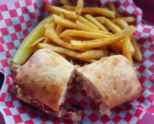 A sandwich cut in half, served with golden fries and a pickle, all on a red and white checkered paper.