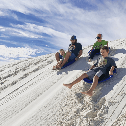Four people sledding down a sandy hill under a blue sky, enjoying a fun day outdoors.