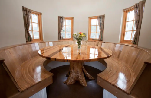 A cozy dining area with a round wooden table and curved wooden benches, surrounded by windows and natural light.
