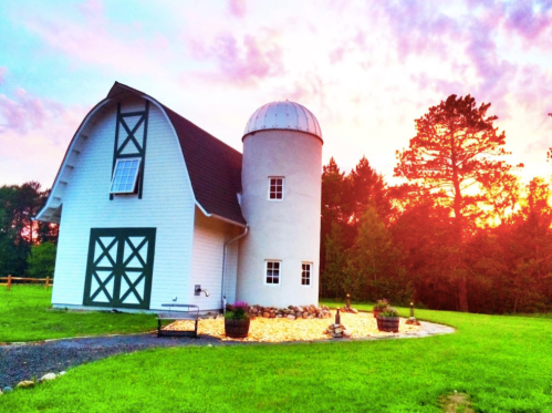 A white barn with a round silo against a vibrant sunset, surrounded by green grass and trees.