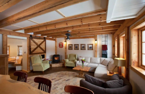 Cozy living room with wooden beams, a fireplace, comfortable seating, and large windows letting in natural light.