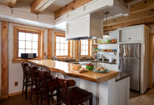 A cozy kitchen with wooden beams, a large island, bar stools, and colorful dishware on shelves.