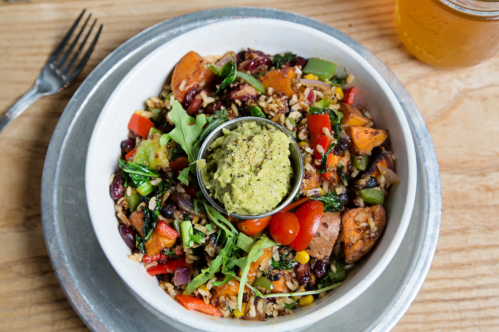 A colorful bowl of mixed vegetables, grains, and a dollop of guacamole, served on a metal tray with a drink nearby.