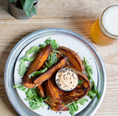 A plate of roasted sweet potatoes with a dipping sauce, garnished with arugula and served with a glass of beer.