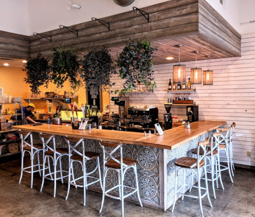 A cozy café interior featuring a wooden bar, hanging plants, and decorative tiles, with seating and a coffee station.