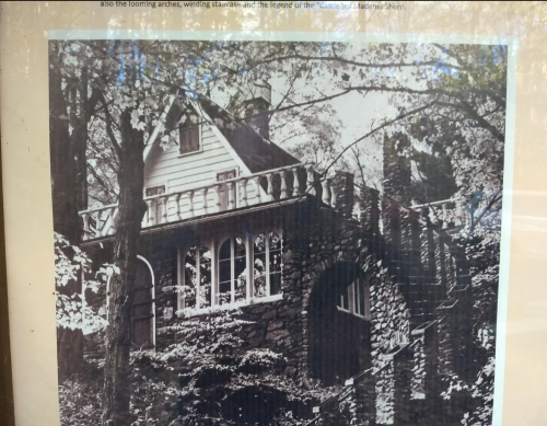 A vintage black-and-white photo of a stone house surrounded by trees, featuring a balcony and winding staircase.