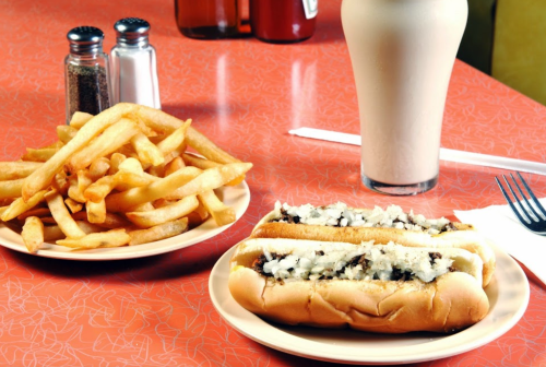 A plate with a hot dog topped with onions, a side of golden fries, and a milkshake on a retro-style table.