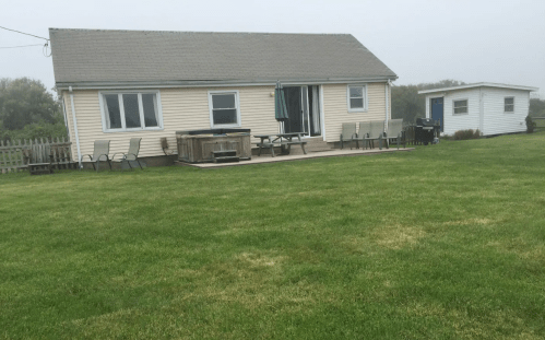 A single-story house with a porch, surrounded by a grassy lawn, in a foggy setting. A small shed is visible nearby.