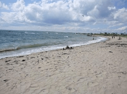 A sandy beach with gentle waves, under a cloudy sky, and a few people enjoying the shoreline in the distance.