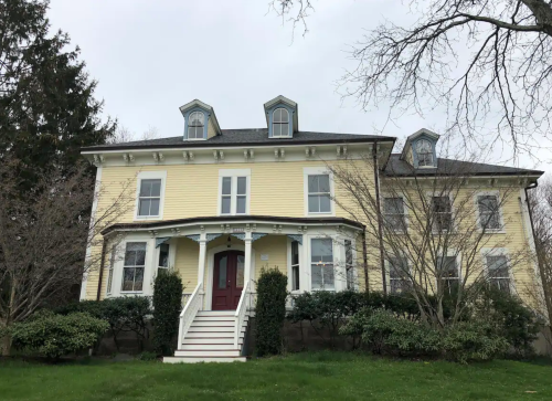 A large yellow Victorian-style house with a front porch and multiple gabled windows, surrounded by greenery.