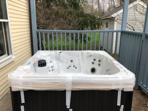 A hot tub wrapped in plastic sits on a deck, surrounded by trees and houses in the background.