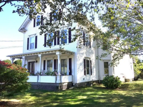 A white two-story house with black shutters, surrounded by greenery and trees on a sunny day.