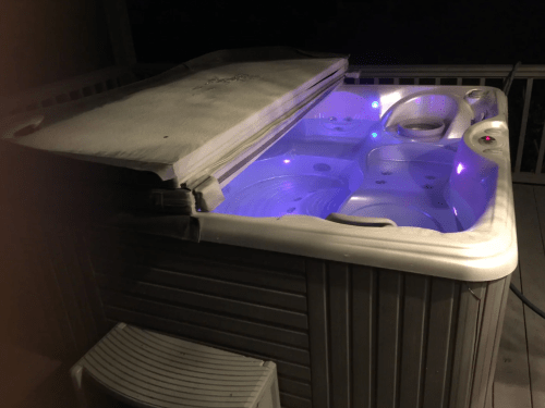 A hot tub with a partially closed cover, illuminated by soft blue lights, set on a deck at night.