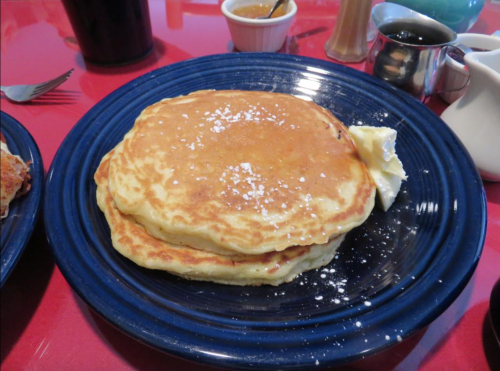 A stack of fluffy pancakes topped with powdered sugar and a pat of butter on a blue plate, with syrup and coffee in the background.