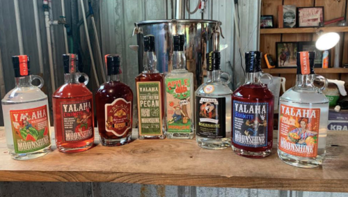 A wooden table displays various bottles of moonshine, featuring colorful labels and different flavors.