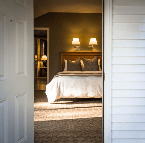 A cozy bedroom with a bed, two lamps, and warm lighting, viewed from an open door.
