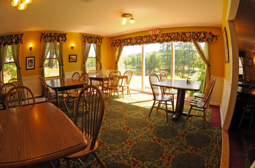 Cozy dining area with wooden tables, chairs, and large windows overlooking a green landscape. Bright and inviting atmosphere.