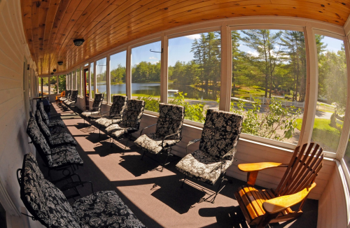 A sunlit porch with patterned chairs overlooking a serene lake and trees in the background.