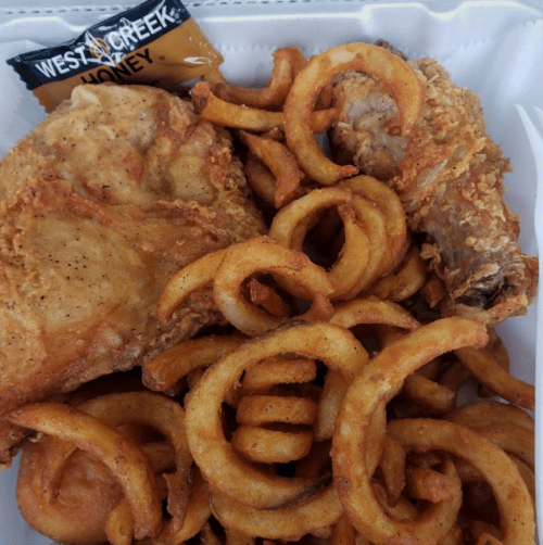 A takeout container with two pieces of fried chicken and a serving of curly fries, accompanied by a packet of honey.