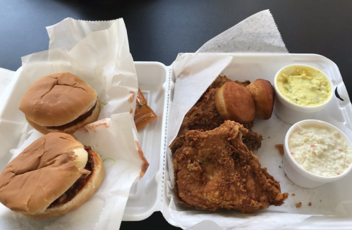 Two sandwiches with sauce, fried chicken, cornbread, and sides of coleslaw and potato salad in a takeout container.