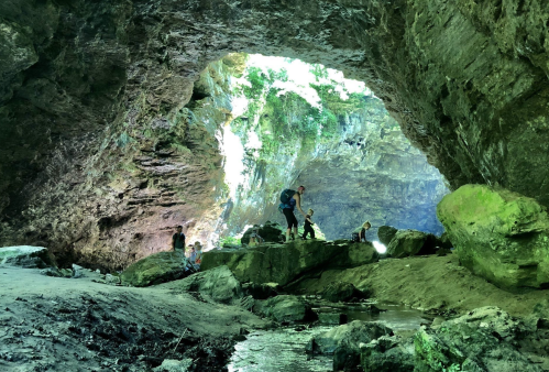 A group of people exploring a large cave with a stream and lush greenery in the background.