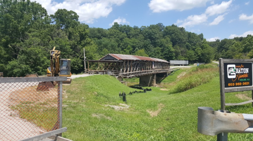 A covered bridge spans a grassy area, with construction equipment nearby and trees in the background under a blue sky.