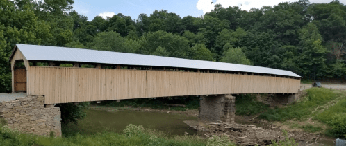 A wooden covered bridge spans a river, surrounded by lush green trees and a clear blue sky.