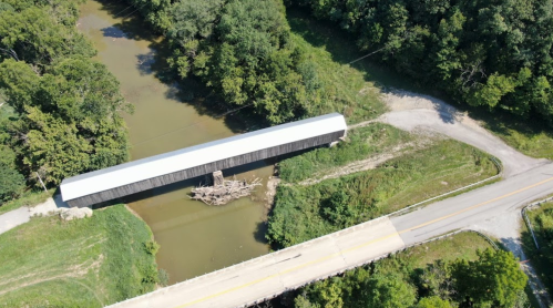 Aerial view of a covered bridge over a river, surrounded by greenery and a nearby road.