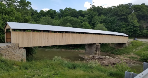 A wooden covered bridge spans a river, surrounded by lush greenery and a clear blue sky.