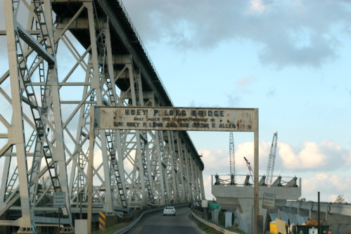 A view of the Huey P. Long Bridge with a weathered sign, surrounded by construction and a cloudy sky.