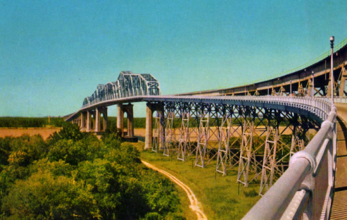 A vintage postcard image of a large, steel bridge arching over green trees and a winding path below.