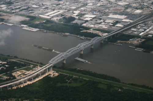 Aerial view of a large bridge spanning a river, with industrial areas and boats visible along the waterway.