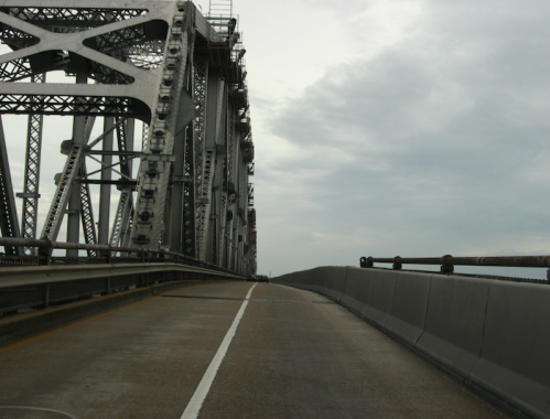 A view of a bridge's structure with a cloudy sky, showing the road and metal framework on one side.