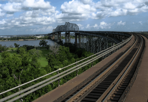 A curved bridge spans over a river, with train tracks running alongside and lush greenery below under a cloudy sky.