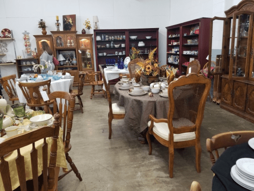 A cozy antique shop filled with wooden furniture, tables set with dishware, and shelves displaying various collectibles.