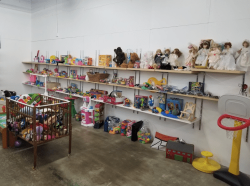 A room filled with shelves of toys, dolls, and games, with a playpen in front and a basketball hoop in the corner.