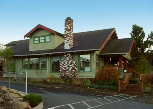 A charming green house with a red roof and a stone chimney, surrounded by trees and a paved parking area.