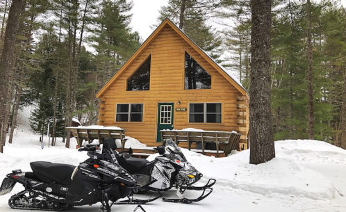 A cozy log cabin surrounded by snow-covered trees, with two snowmobiles parked in front.