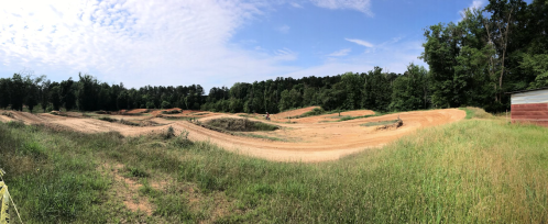 A panoramic view of a dirt bike track surrounded by green grass and trees under a blue sky with scattered clouds.