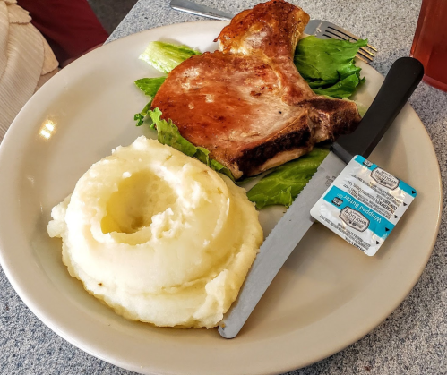 A plate with a grilled pork chop, lettuce, mashed potatoes, and a packet of butter on a gray table.