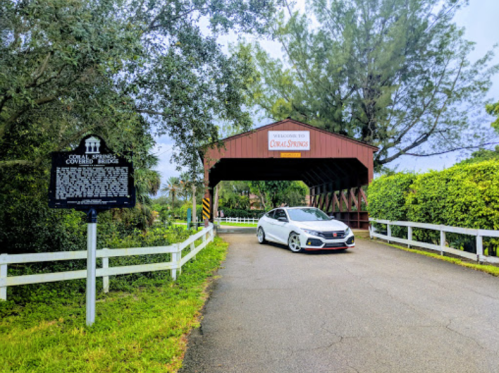 A white car drives through a covered bridge, with a historical sign and lush greenery surrounding the entrance.