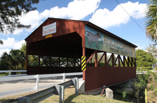 A red covered bridge with a sign reading "Bull of the Woods" surrounded by trees and blue sky.
