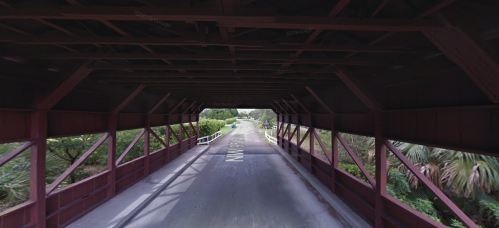 A view from inside a red covered bridge, looking out onto a road surrounded by greenery.